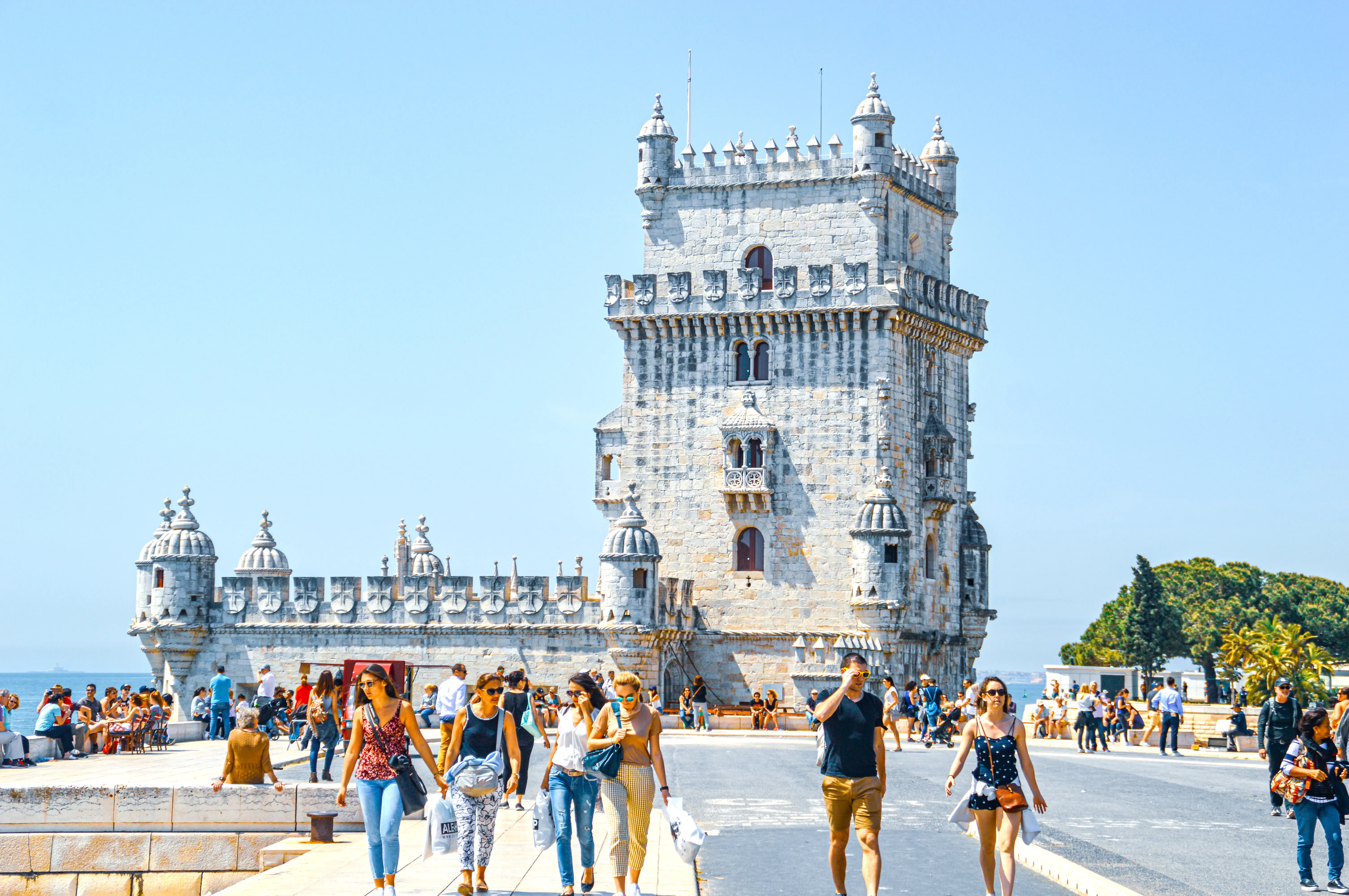 Tourists walk along the waterfront in front of the historic Belém Tower in Lisbon on a bright, sunny day.